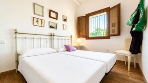 A double bed with white linens, a bedside table with a lamp, and a chair with a green bag.