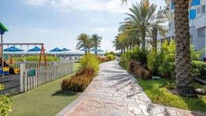 Un vialetto alberato da palme e un’area giochi con ombrelloni azzurri.