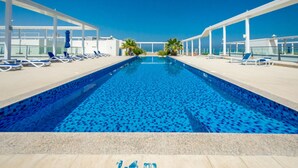 Una piscina con piastrelle di mosaico blu, circondata da poltroncine bianche e ombrelloni, con un cielo azzurro limpido sullo sfondo.