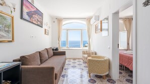 Un salon comprenant un canapé, des fauteuils, une table à manger et offrant une vue sur la mer grâce à une grande fenêtre.