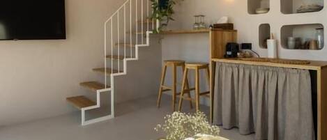 A modern living space with a staircase, a flat-screen TV, a coffee table with books and a vase, and a wall-mounted shelf with decorative items.