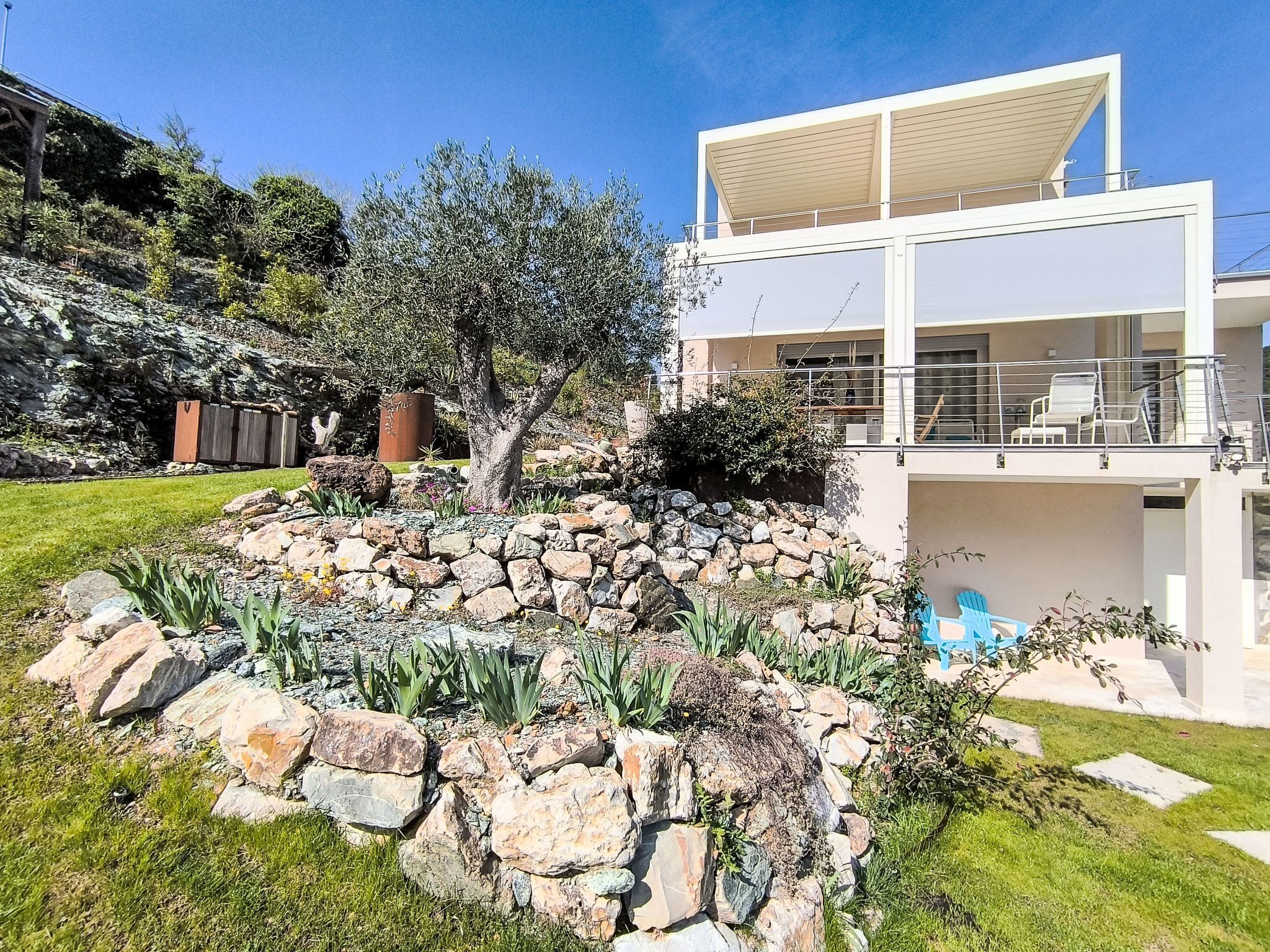 A modern two-story house with a stone garden, a tree, and a clear blue sky.