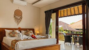 A bedroom with a wooden bed, a headboard, a wall-mounted air conditioner, a dreamcatcher, and a balcony with chairs and a table. - Putu's Paradise Ubud (Ubud)