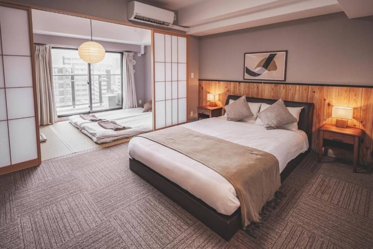 A hotel room with a large bed, two bedside tables with lamps, a sliding shoji screen door, and a city view through the window.