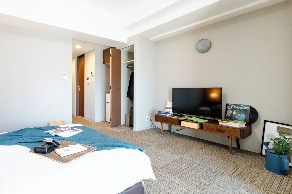 A hotel room with a bed, a desk with a TV, a clock, and a plant.