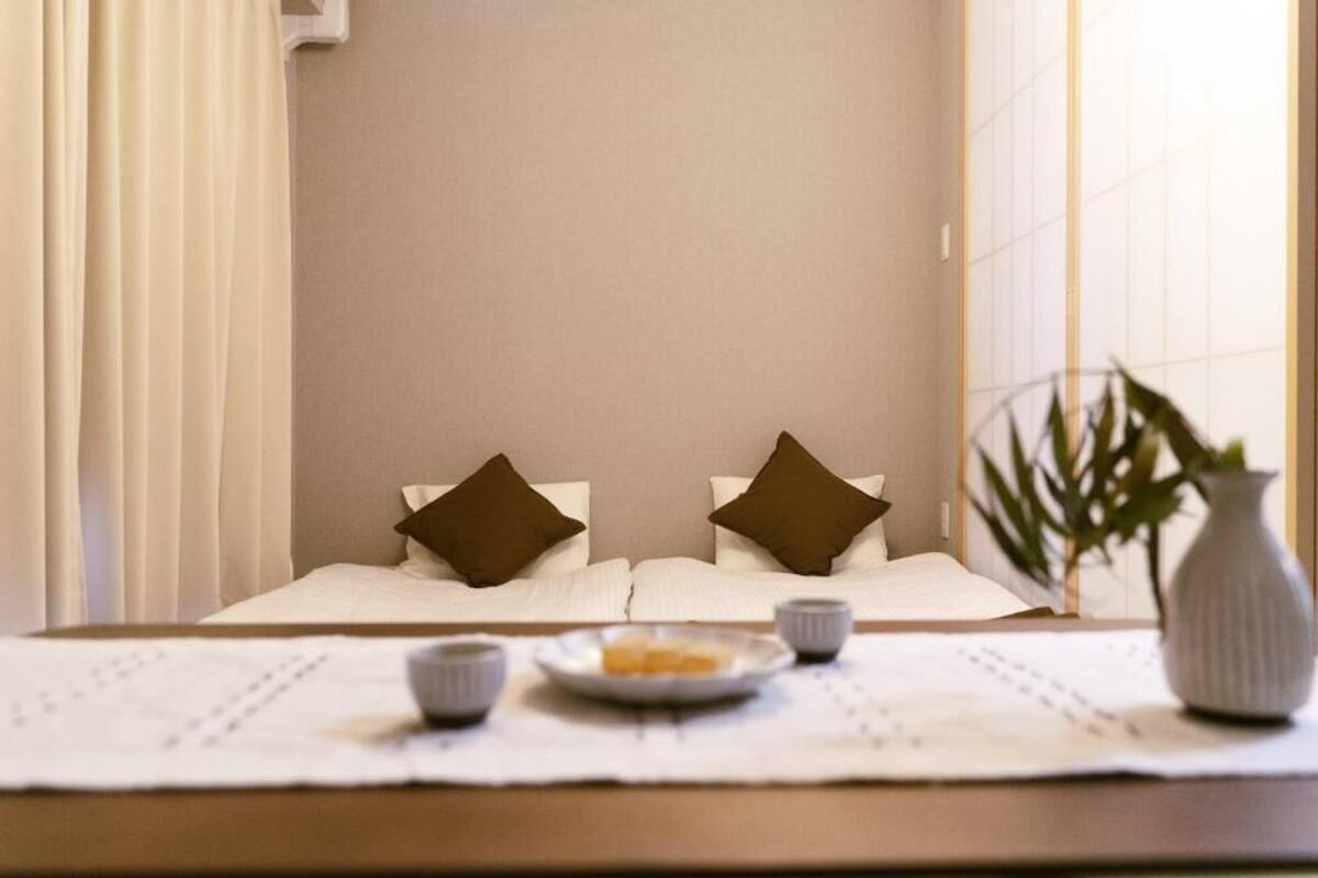 A neatly made bed with two pillows, a small table with a plate of food and two cups, and a vase with a plant.