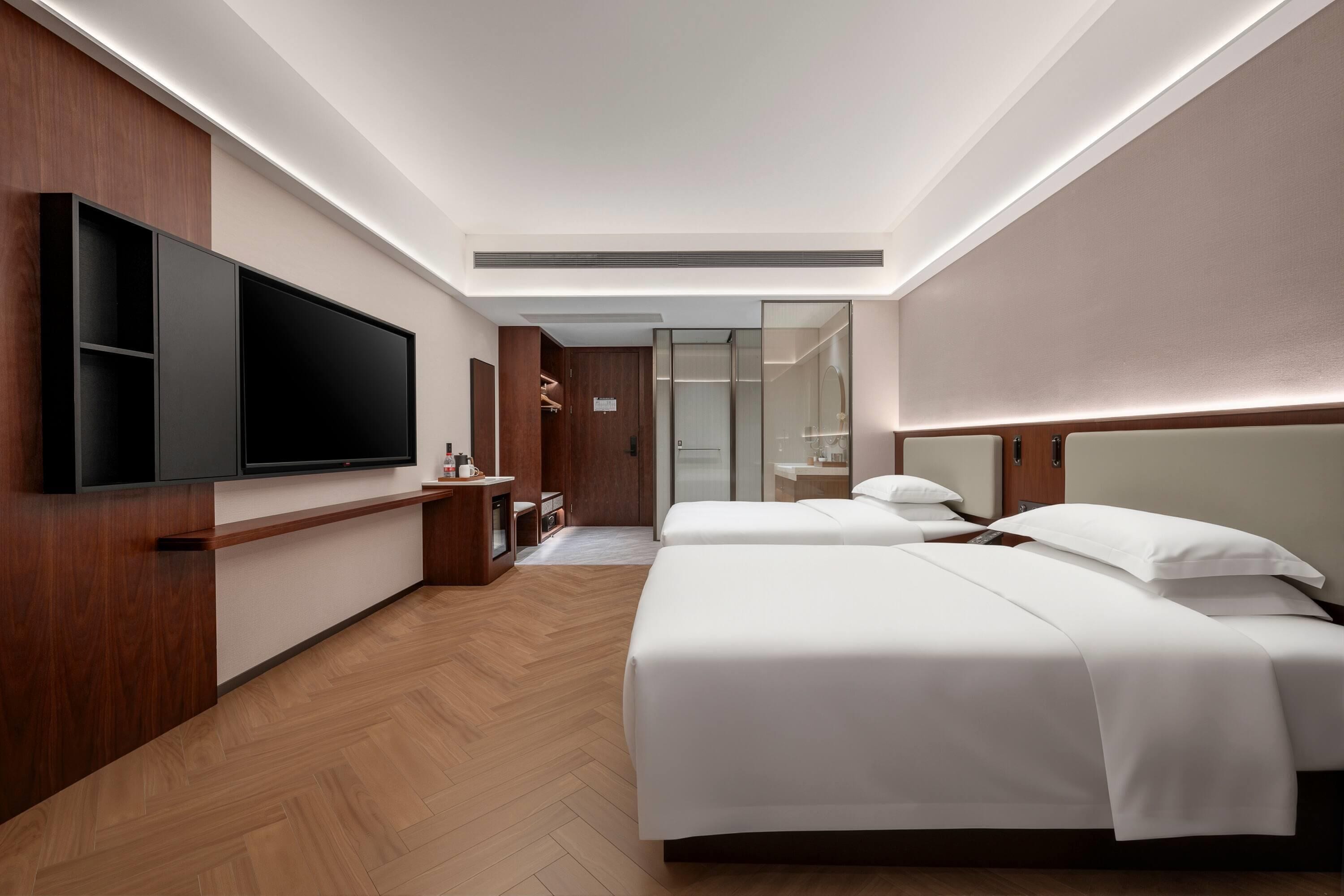 A hotel room with a large flat-screen TV mounted on the wall, a wooden wardrobe, a bedside table with a lamp, and a bed with white bedding.