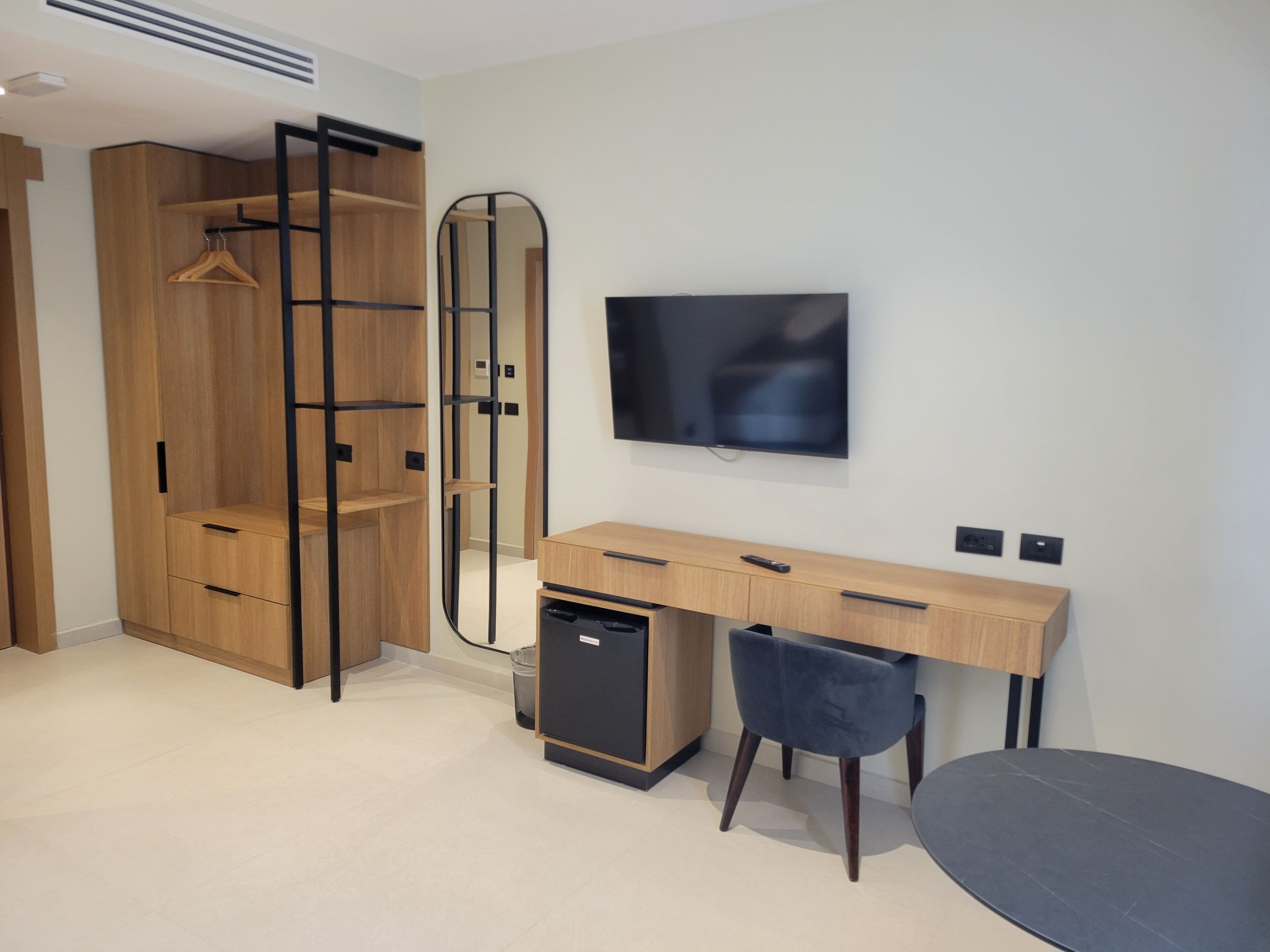 A modern room with a wooden wardrobe, a desk with a chair, a television, and a small trash bin.