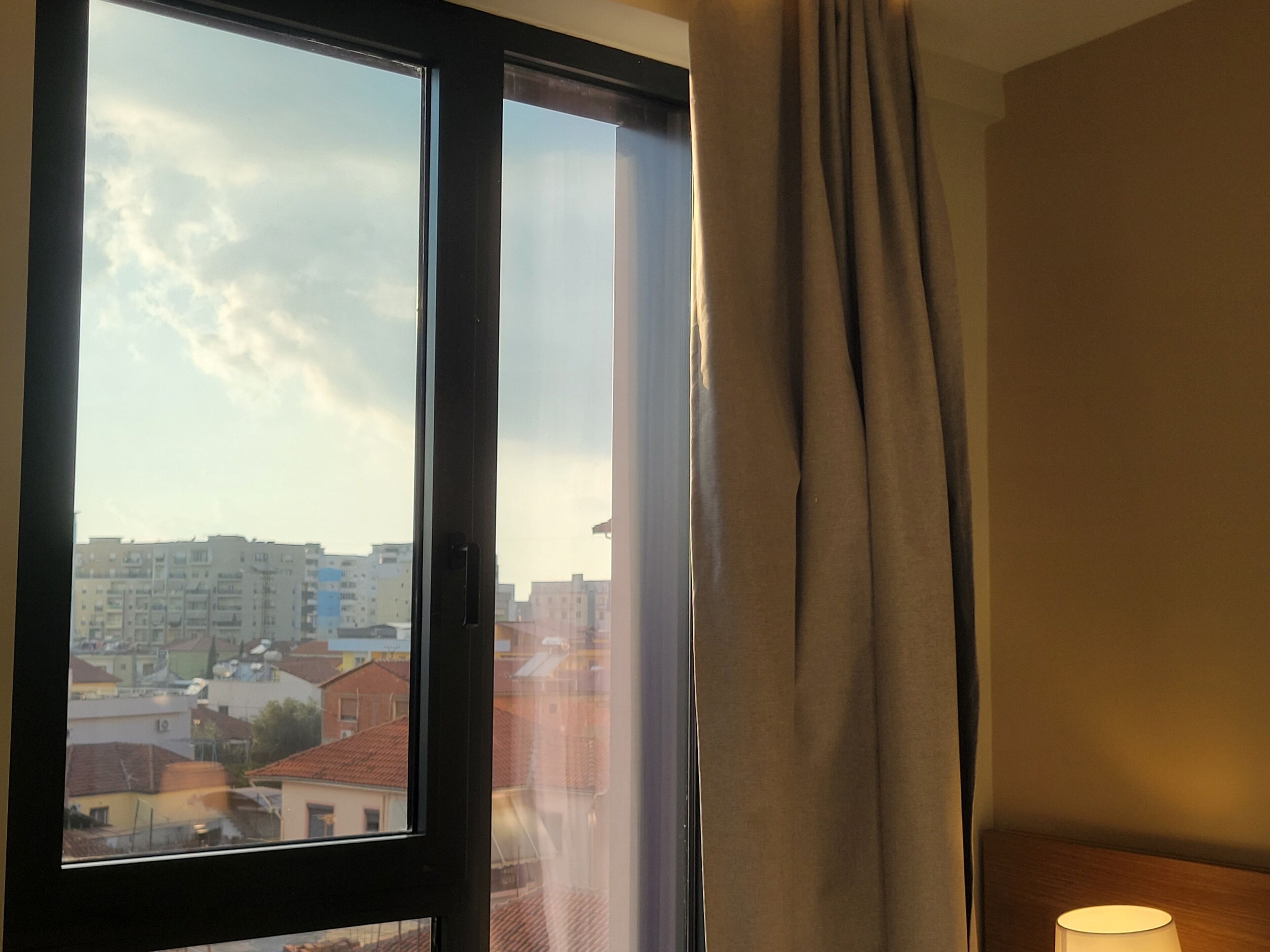 Una habitación con un ventanal grande, una cortina y una lámpara sobre una mesita de noche.