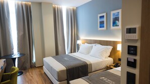 Quarto de hotel com duas camas, uma mesa pequena, uma cadeira e mesa de cabeceira com abajur.