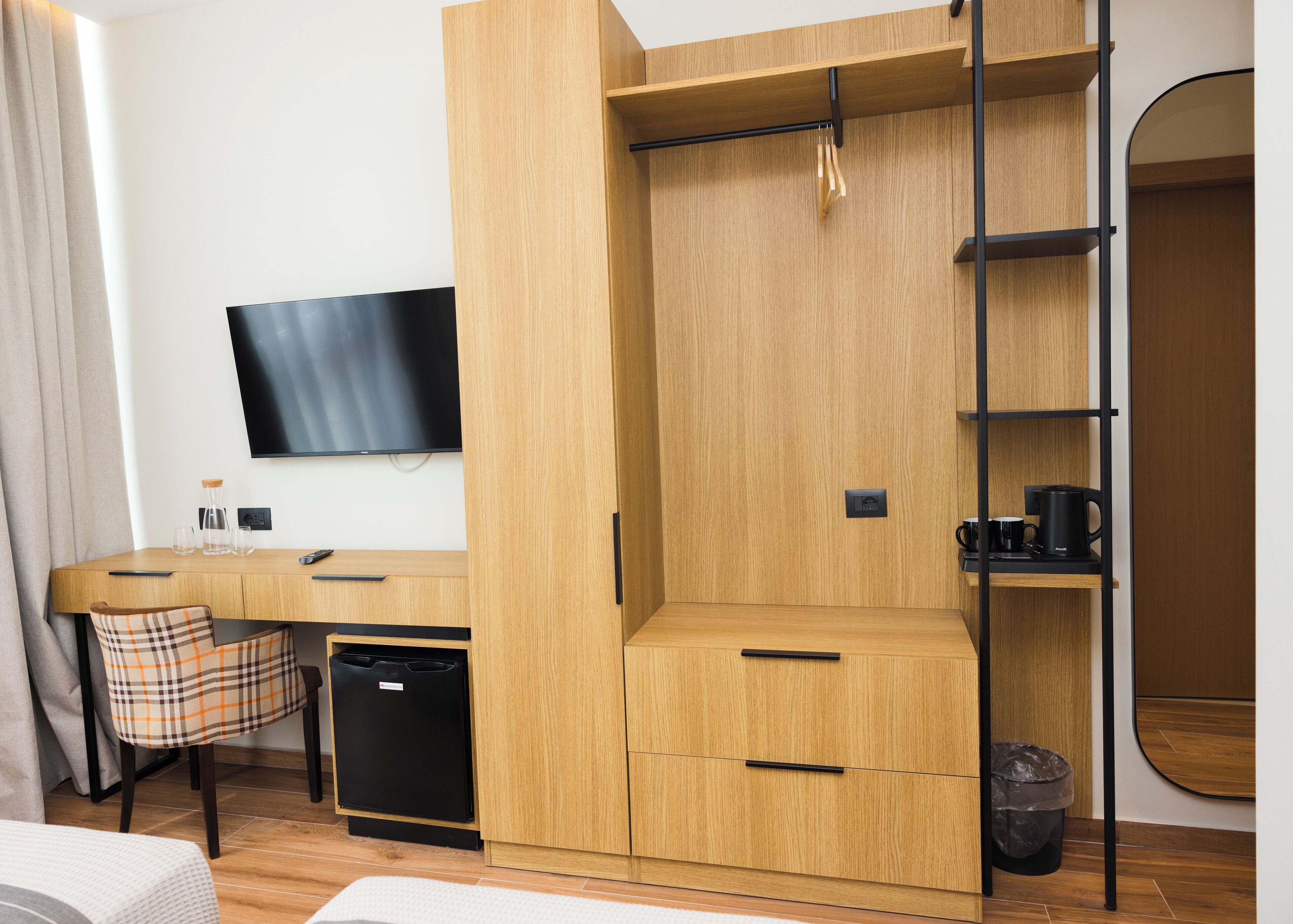 Quarto de hotel compacto com guarda-roupa de madeira, uma escrivaninha com cadeira, televisão e um pequeno cesto de lixo.