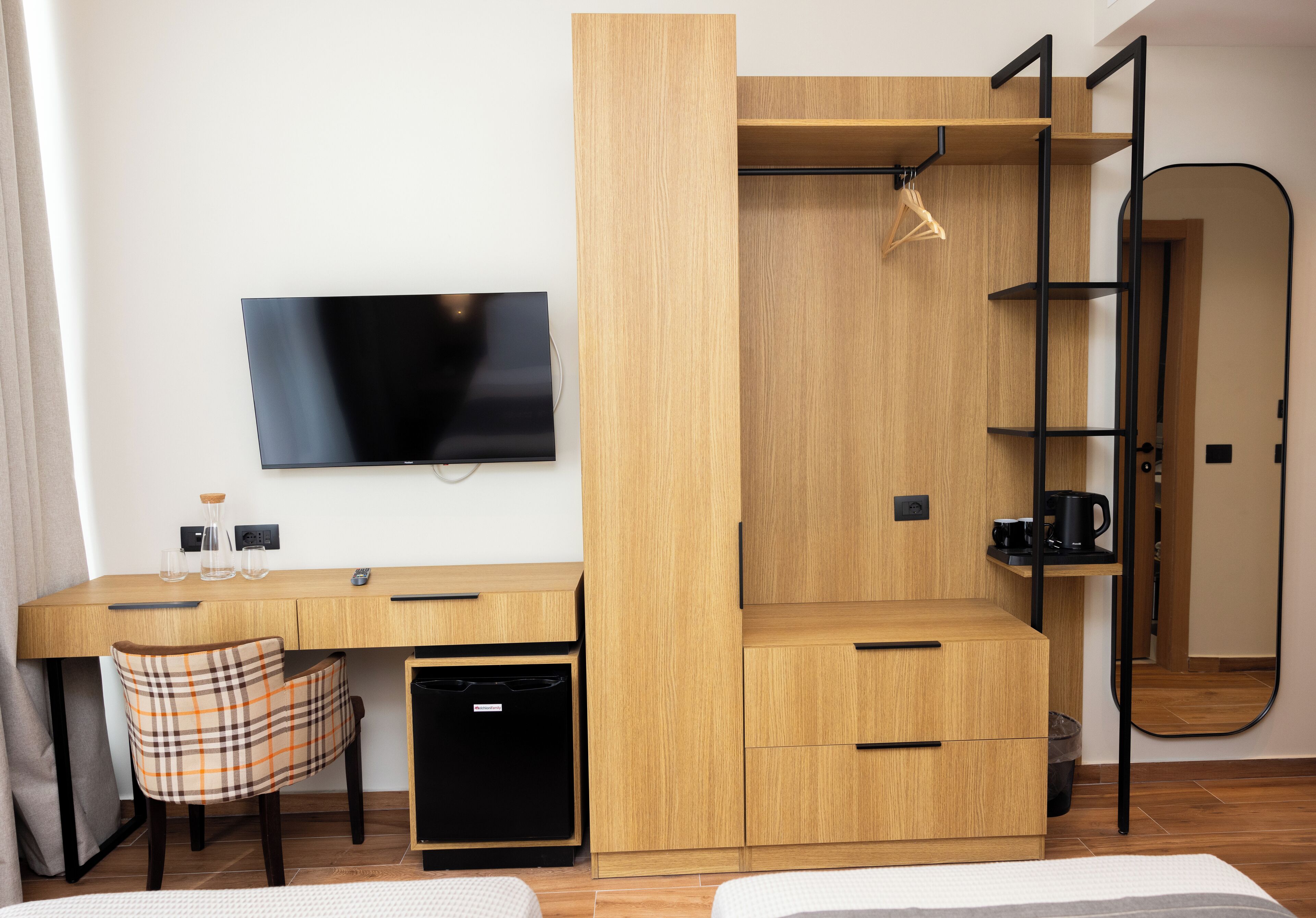 A compact room with a wooden desk, a chair, a television, a wardrobe, and a mirror.