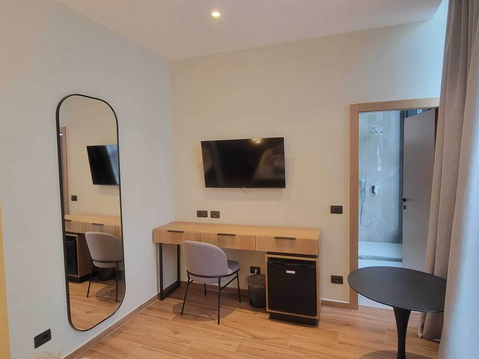 A modern hotel room with a wooden desk, a flat-screen TV, a mirror, a small black refrigerator, a chair, and a shower area.