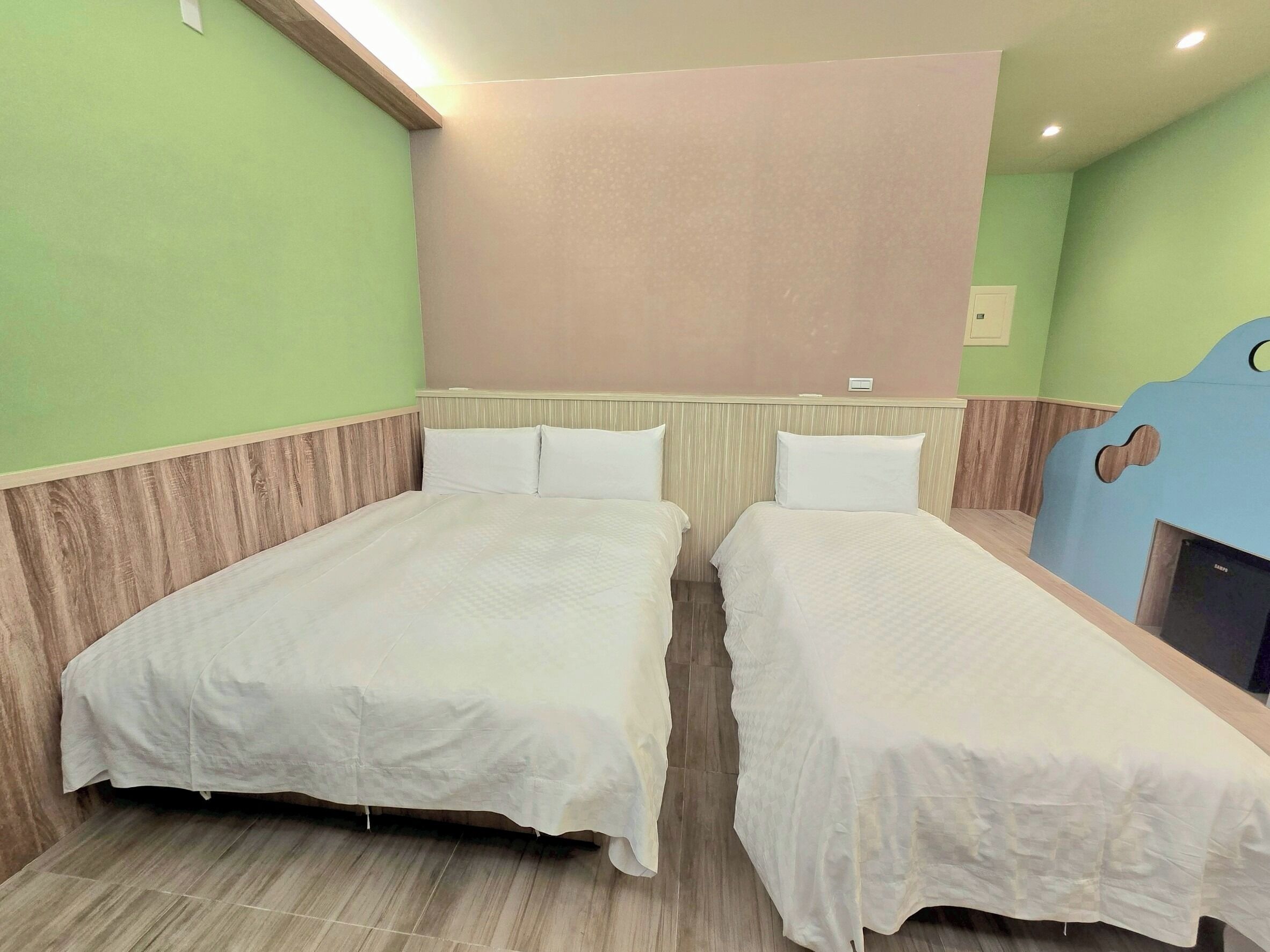 Une chambre d’hôtel avec deux lits simples, une tête de lit en bois, un mur de couleur claire et un petit téléviseur.