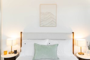 A hotel room with a bed, two bedside tables with lamps, a nightstand with a lamp, a wall-mounted artwork, and a small plant. - Chada by Cadiz Properties – Lakeview | Pool | CBD & Parkside (Canberra)