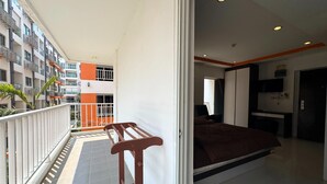 A balcony with a wooden bench, a bed with a brown bedspread, and a view of a multi-story building with orange and white colors. - Stylish Pool-view Studio With Balcony - New Nordic VIP 3, Pratumnak (Pattaya)