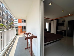 Ein Balkon mit einer hölzernen Bank, einem Bett mit brauner Tagesdecke und Blick auf ein mehrgeschossiges Gebäude in Orange und Weiß.