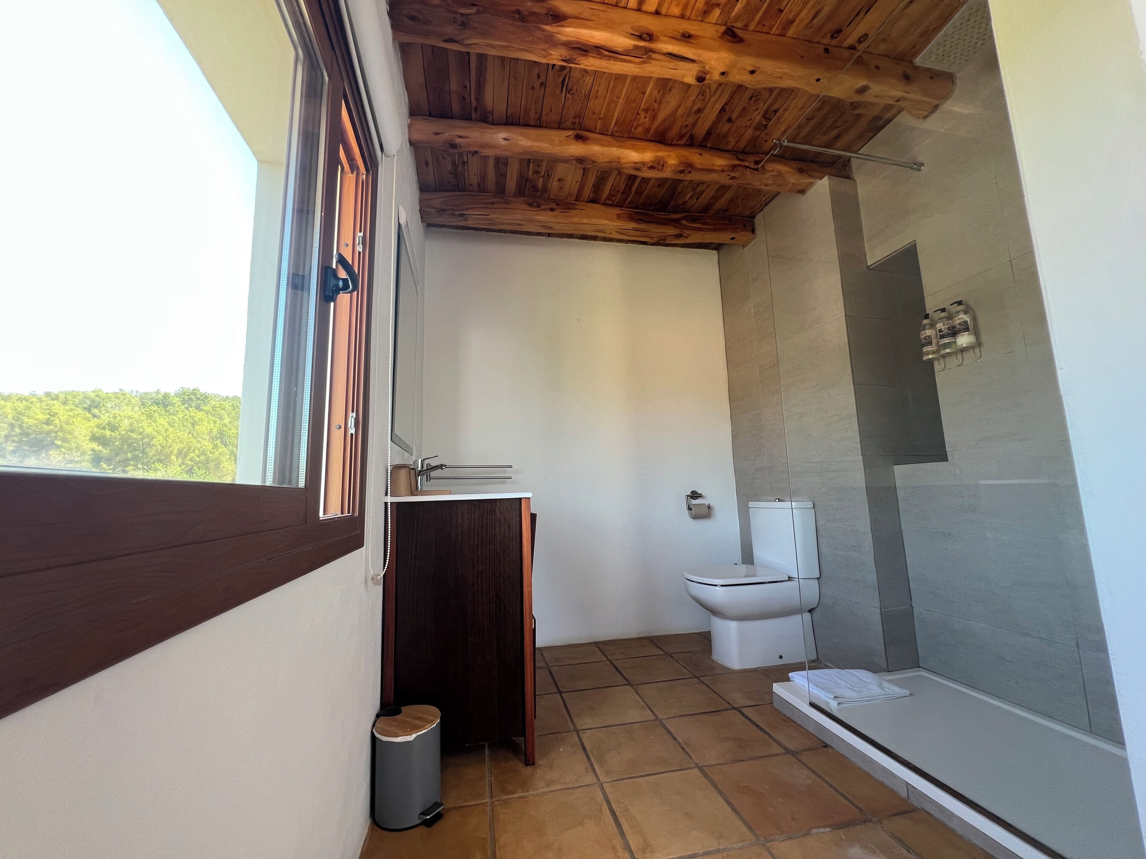 A bathroom with a wooden ceiling, a toilet, a sink, and a trash bin.