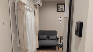 Un couloir mène à une pièce où se trouvent un canapé gris, un fauteuil et une paire de pantoufles posées sur le sol.