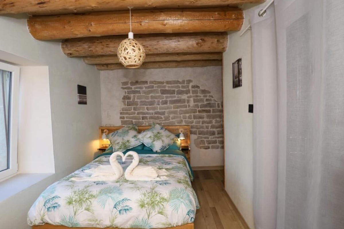 Een slaapkamer met een bed, houten balken in het plafond, een stenen muur en een raam.