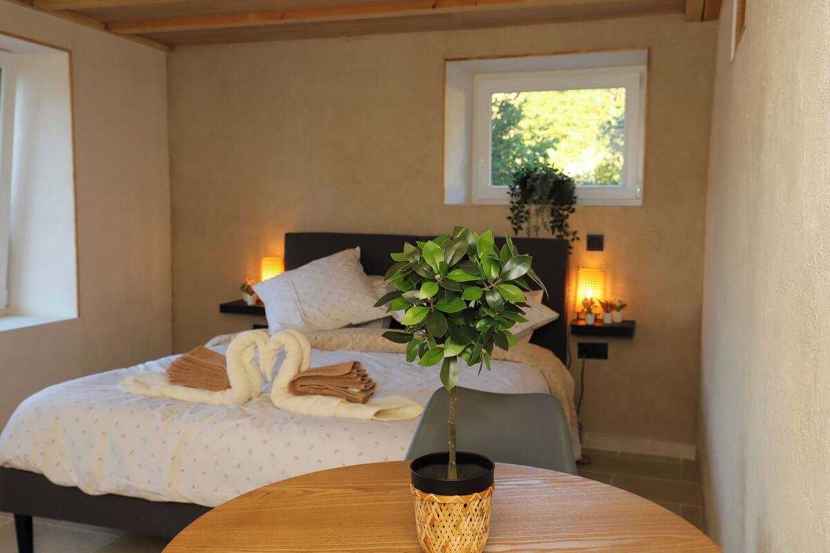 Een slaapkamer met een net opgemaakt bed, een nachtkastje met een lamp en een raam met uitzicht op bomen.