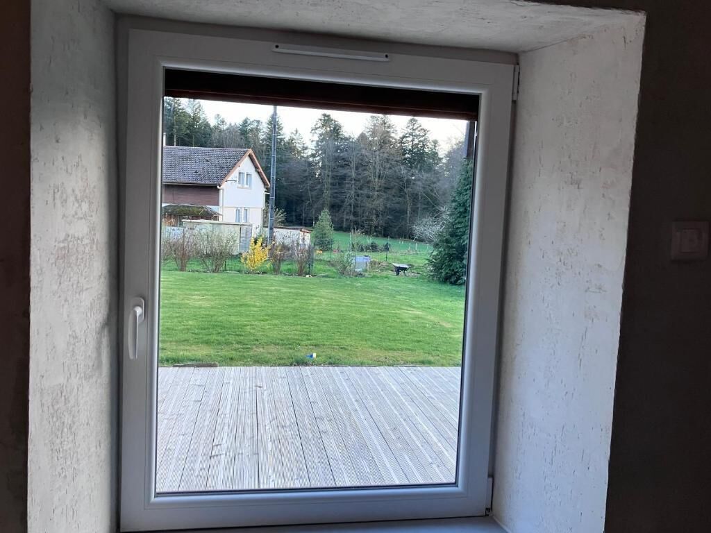 Une pièce dotée d’une grande fenêtre donnant sur une terrasse en bois et une cour verdoyante avec des arbres.