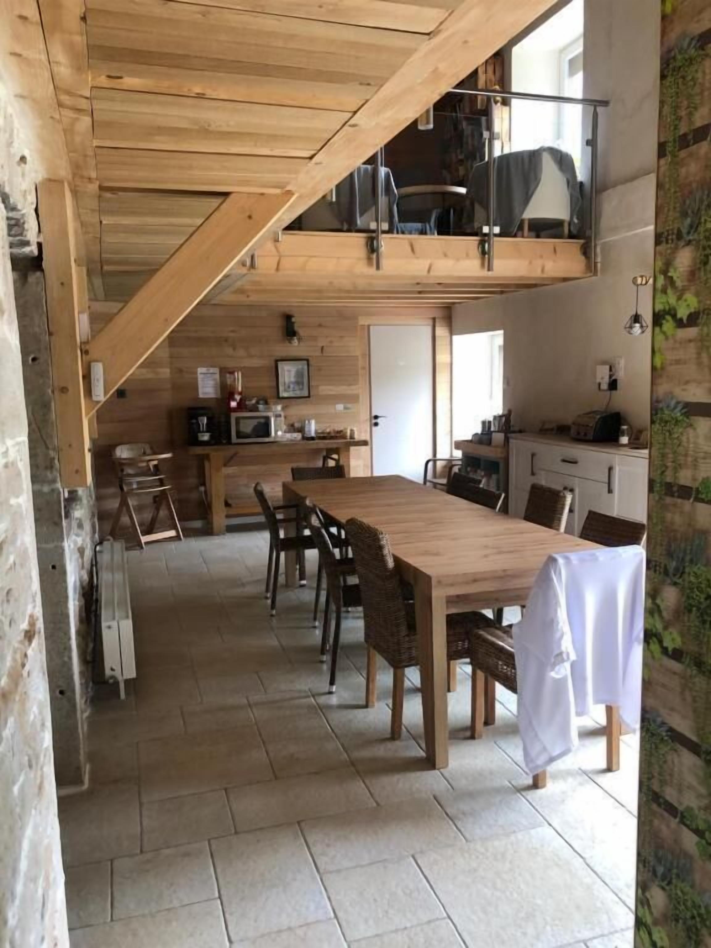 Un grand espace à manger avec une longue table et des chaises en bois, un coin cuisine équipé d’un four à micro-ondes et de placards, et un espace en mezzanine accessible par un escalier.