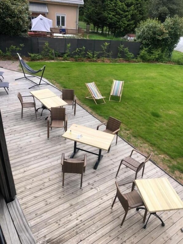 A wooden deck with outdoor furniture including tables and chairs, a hammock, and a grassy yard with a fence and trees.