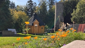 Une cabane en bois, dotée d’un toit en pente, entourée d’un jardin où poussent des coquelicots orange et d’autres fleurs sauvages.