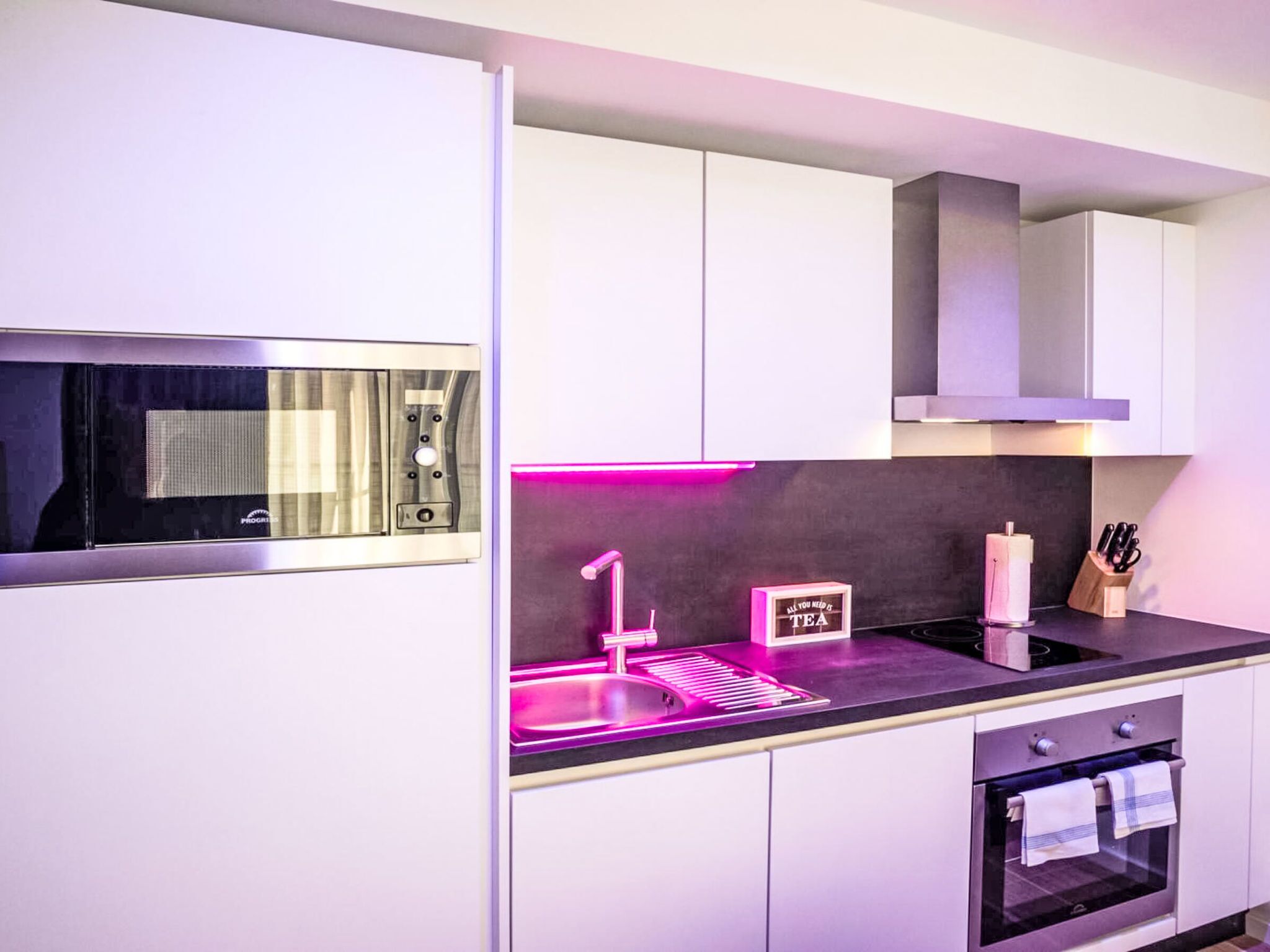 Eine moderne Küche mit integriertem Mikrowellenherd, einer Spüle mit Neonbeleuchtung, einem Schild mit der Aufschrift 'ALL YOU NEED IS TEA' und einem Backofen.