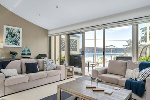 A modern living room with two sofas, a coffee table, and a large sliding glass door leading to a balcony with a view of the sea. - 3-Bed with Beach Views and Entertainment Area (Denhams Beach)