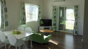 A spacious living room with a wooden ceiling, a dining area with white chairs, a green sofa, and a television. - Yukura Nakijin (Nakijin)