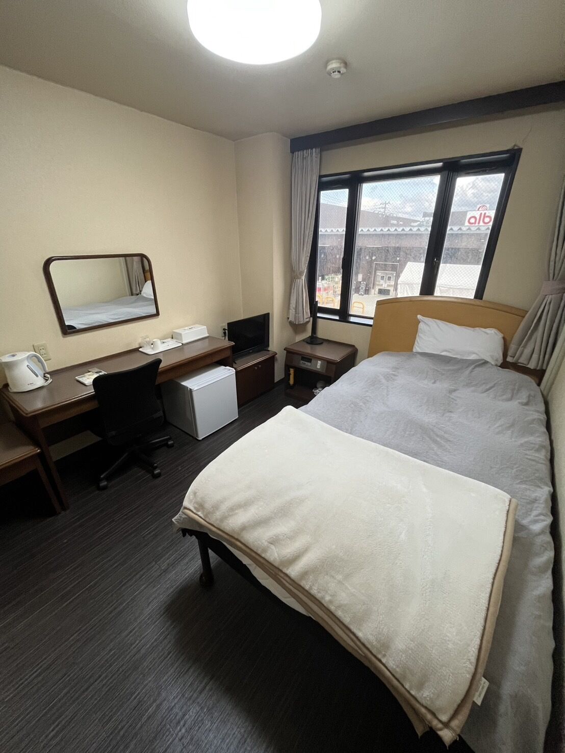 A hotel room with a bed, a desk, a chair, a television, a window with curtains, and a ceiling light.