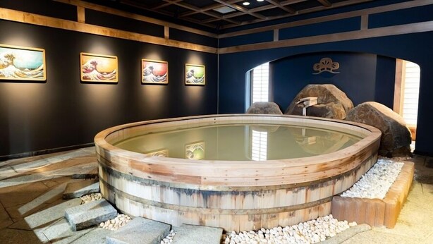 A traditional Japanese onsen with a wooden tub, stone steps, and artwork on the walls. - Sumie Chita (Minamichita)