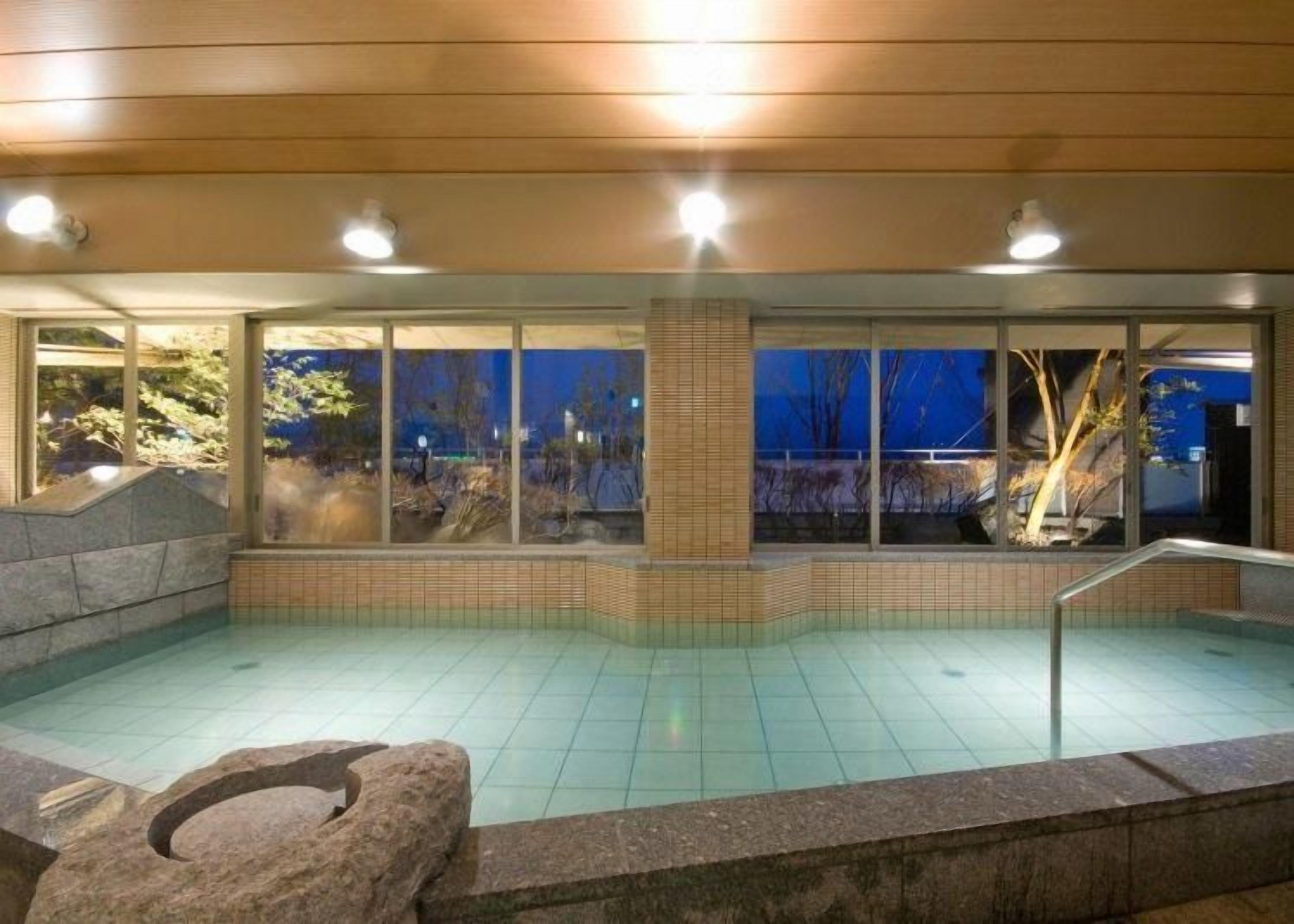 A swimming pool with a stone border, a stone bench, and a view of trees through large windows.