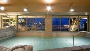 A swimming pool with a stone border, a stone bench, and a view of trees through large windows.