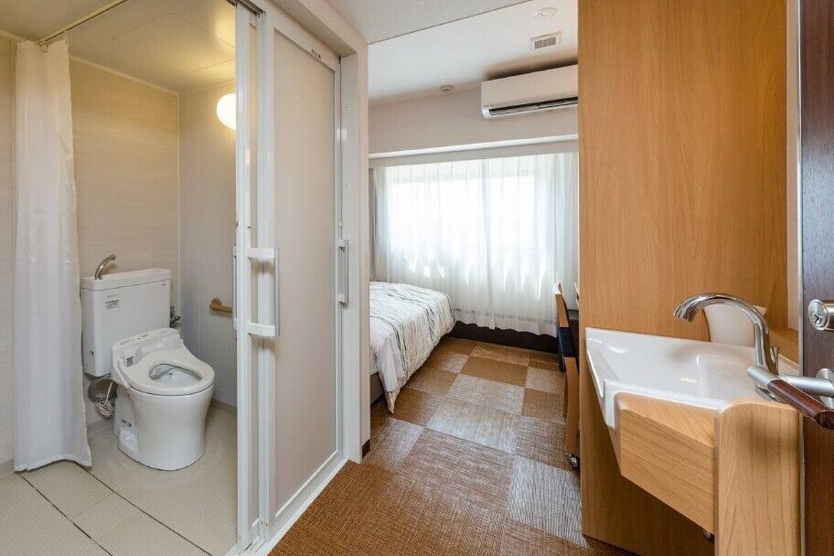 Un baño con inodoro, ducha y lavamanos, una cama en la habitación contigua y un equipo de aire acondicionado en la pared.