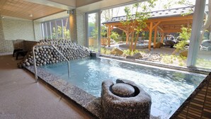 An indoor hot spring area with a stone wall, a wooden deck, and a view of the surrounding garden. - Manten no Yu Michi no Yado (Hakusan)