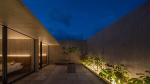 Un espace extérieur moderne avec un mur en béton, un espace clos par des parois de verre contenant un lit et une plante en pot.