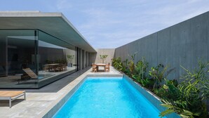 Un espace piscine extérieur moderne, doté d’un espace repas clos par des parois de verre et d’un mur en béton.