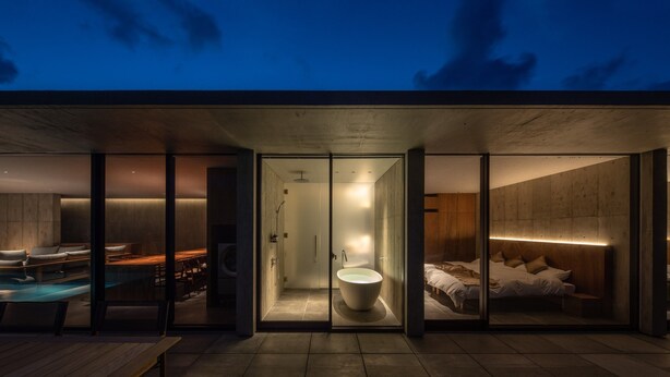 Un espace de vie moderne à aire ouverte, doté d’une salle de bain fermée par des parois de verre, d’un espace piscine et d’une chambre avec un grand lit et un éclairage intégré dans la tête de lit.