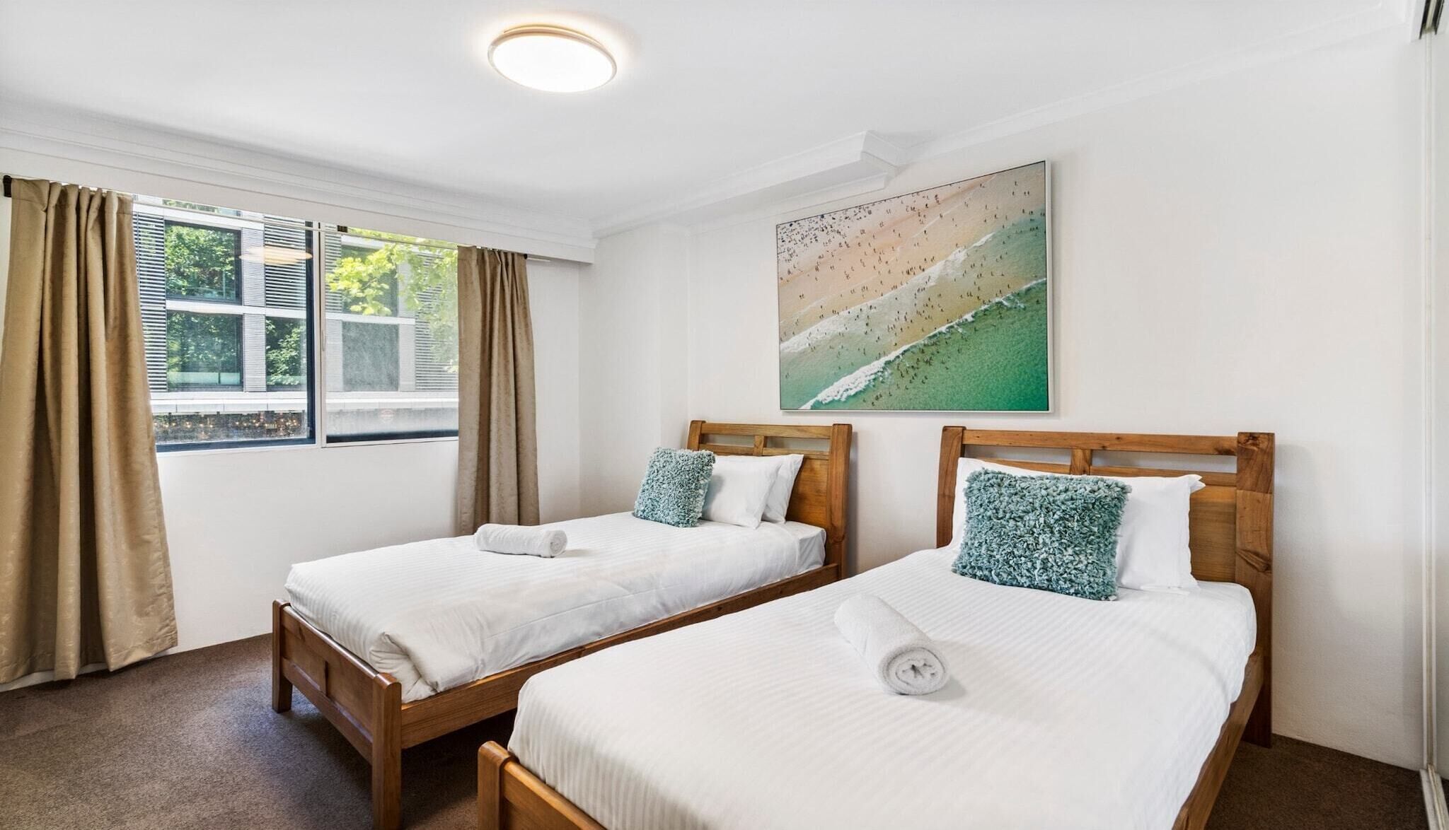 A room with two wooden beds, beige curtains, a window with blinds, and a beach-themed painting on the wall.