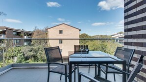 Un balcón con mesa y sillas de vidrio, dos copas de vino y vista a edificios y árboles cercanos.
