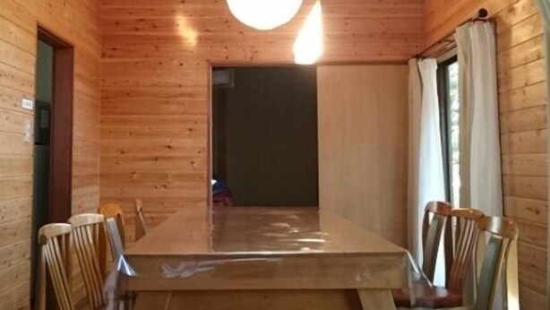Un intérieur en bois avec une table et des chaises au centre, un luminaire suspendu et une porte donnant sur une autre pièce.