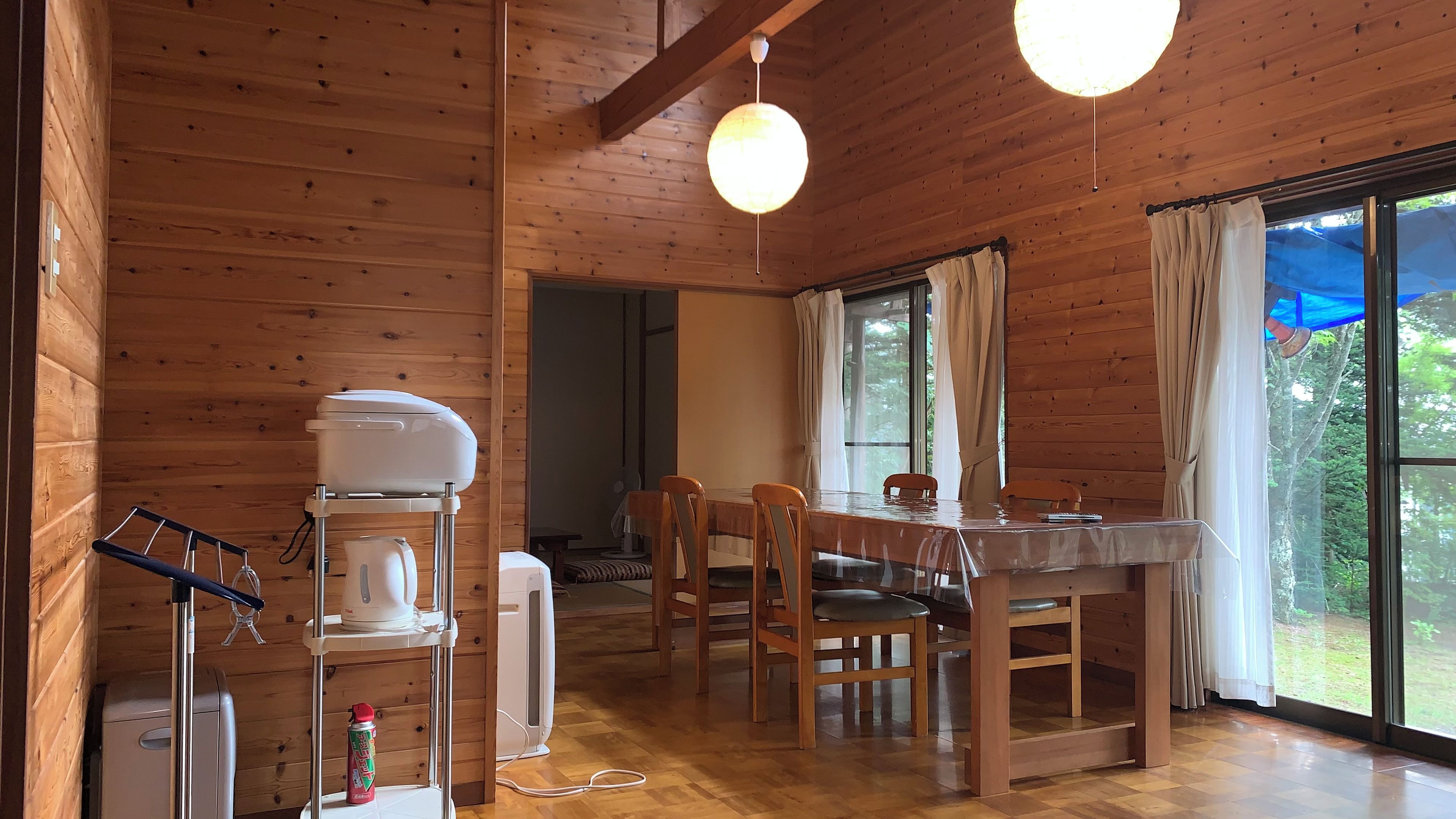 A wooden cabin interior with a dining table, chairs, a coffee machine, and a large window with curtains.
