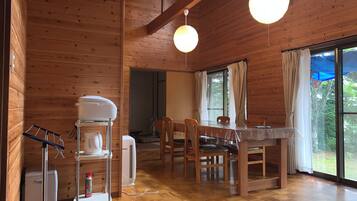 A wooden cabin interior with a dining table, chairs, a coffee machine, and a large window with curtains.