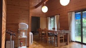 Interior de una cabaña de madera con una mesa de comedor, sillas, una cafetera y un ventanal con cortinas.