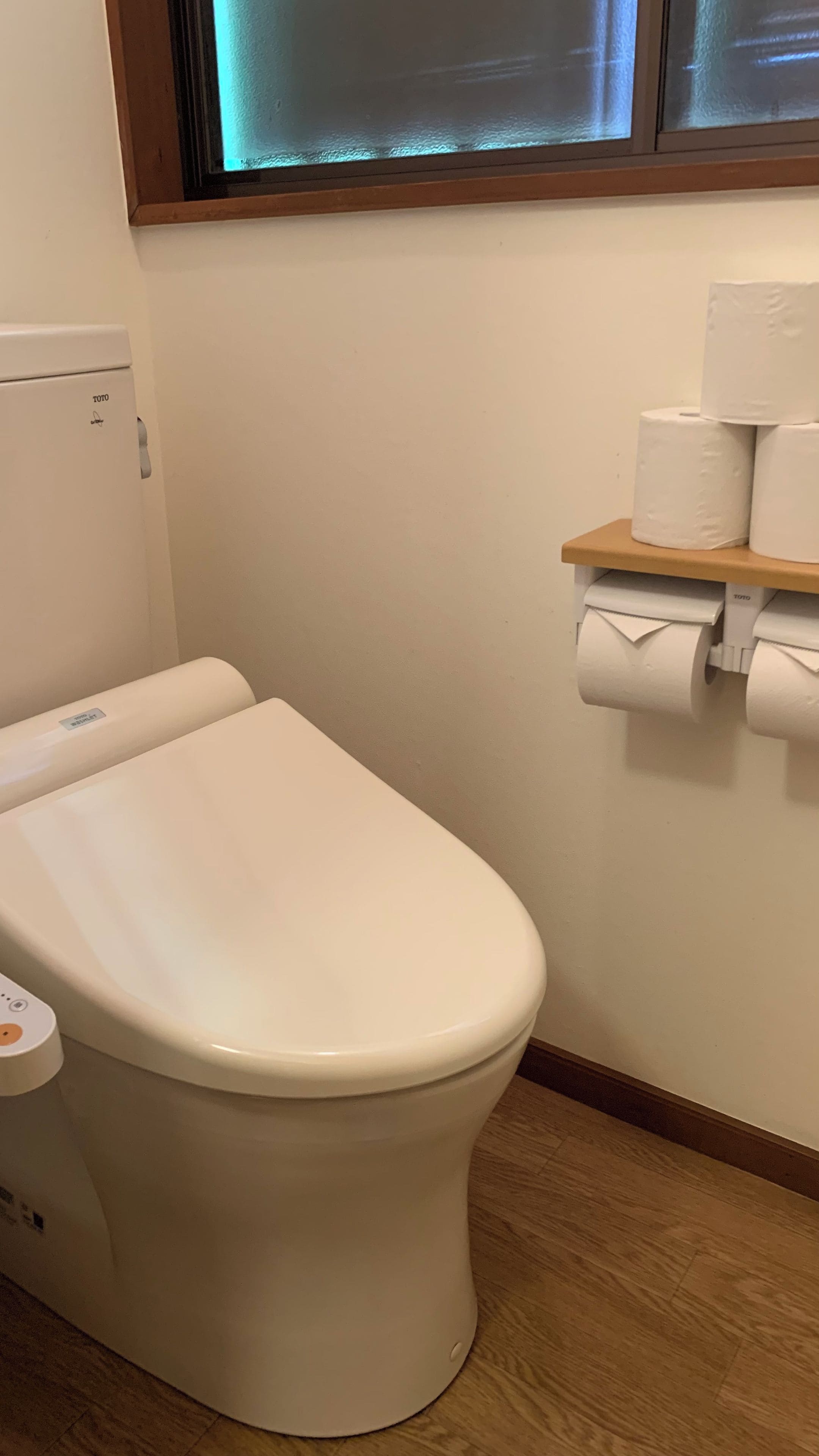A toilet with a TOTO brand label, a toilet paper holder with rolls, and a window above the toilet.
