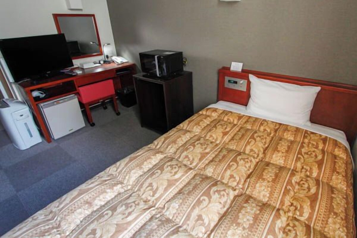 Una habitación de hotel con cama, escritorio, televisor, refrigerador pequeño y mesita de noche.