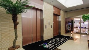 Vestíbulo de hotel con mostrador de recepción, una planta en maceta y alfombra negra con el nombre del hotel.